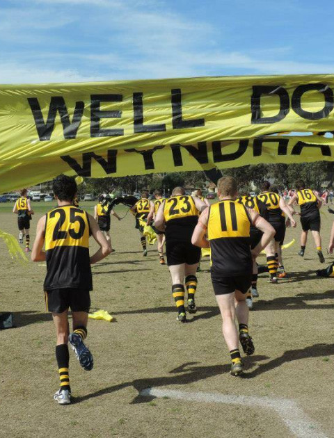 a group of men wearing yellow and black sports clothing run under a yellow banner saying Well Done