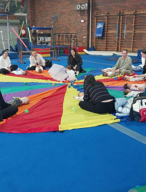 a group of adults sit around a parachute whilst babies crawl ontop