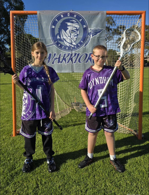 a boy and girl in purple and black sports uniforms stand infront of a goal holding Lacrosse sticks