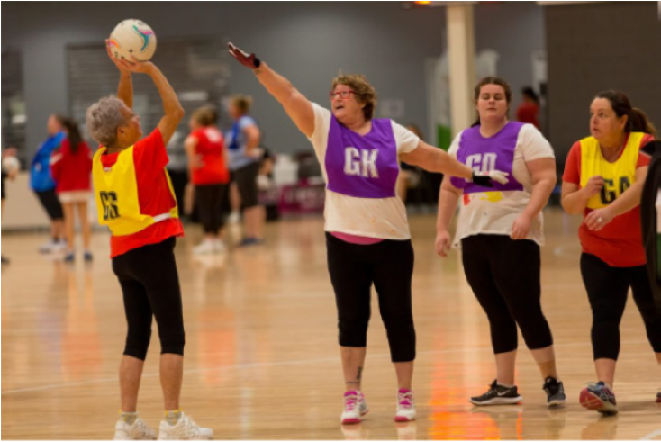 A woman throws a ball towards netball post while three other women stand with their arms up between her and the Netball post