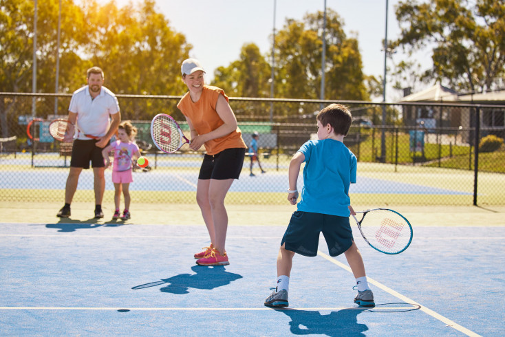 a child dressed in blue is swinging a Tennis racquet at a tennis ball, whilst 2 adults and a child watch