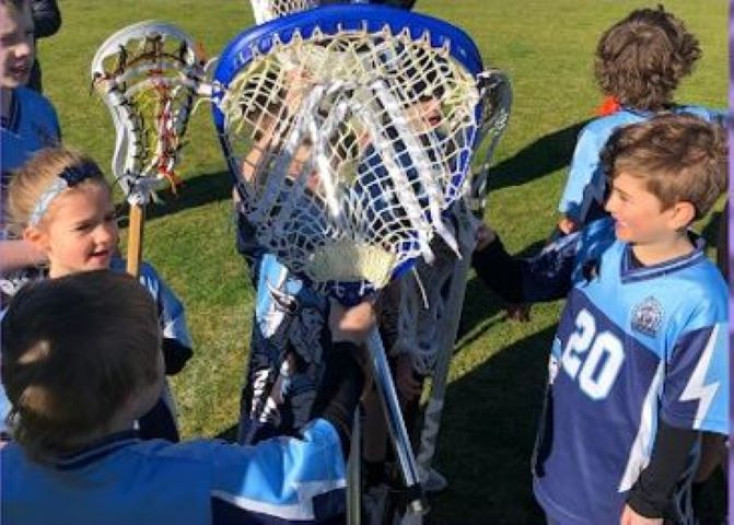 children in a circle are standing around a lacrosse stick held up to the camera