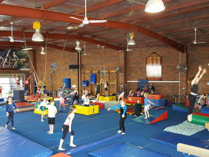 children are spread around different activity stations on blue mats, one is jumping, another has a hoop, another is balancing on a blue beam, another is trying a handstand