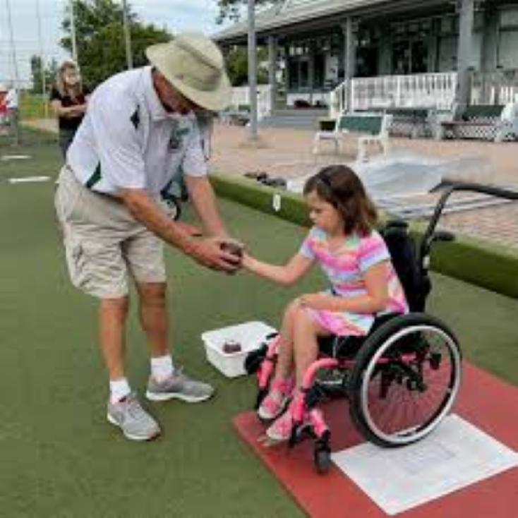 a young girl in a wheelchair being handed a bowling ball from a bowling coach on the green