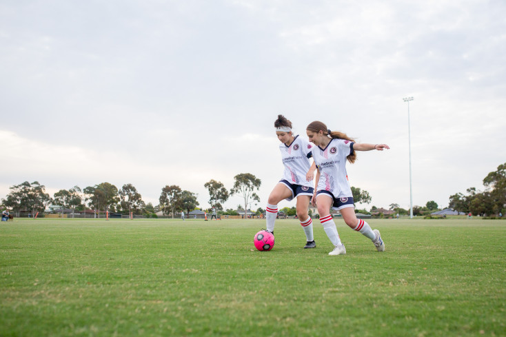 two girls in white and blue sports uniforms are running after a pink football