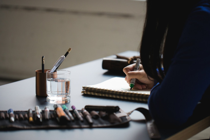 Side view of girl writing calligraphy at a desk with lots of coloured pens around her.