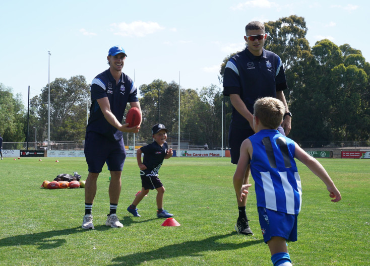 2 men are smiling at 2 children who are running between cones on a grass pitch