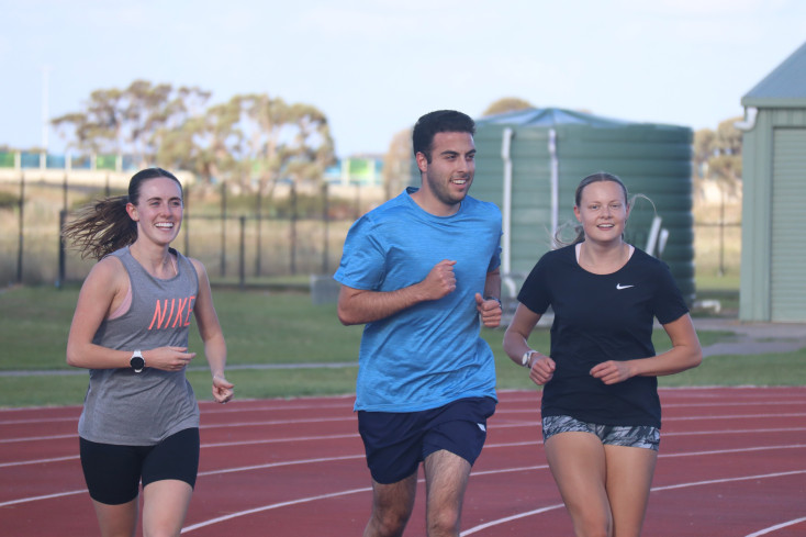 3 people wearing sports clothes are jogging around an athletics track, smiling at the camera