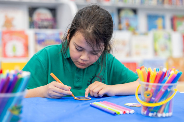 A girl sits at a table colouring in a small piece of wood, surrounded by coloured textas.