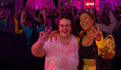 Two ladies are smiling at the camera in a dark room with disco lights and a crowd in the background