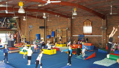 children are spread around different activity stations on blue mats, one is jumping, another has a hoop, another is balancing on a blue beam, another is trying a handstand