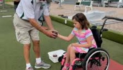 a young girl in a wheelchair being handed a bowling ball from a bowling coach on the green