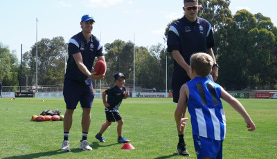 2 men are smiling at 2 children who are running between cones on a grass pitch