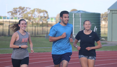 3 people wearing sports clothes are jogging around an athletics track, smiling at the camera