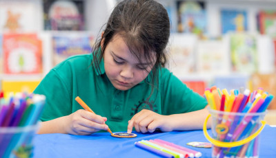 A girl sits at a table colouring in a small piece of wood, surrounded by coloured textas.