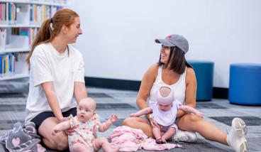 Two mothers and babies sitting in the library and smiling.