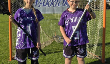 a boy and girl in purple and black sports uniforms stand infront of a goal holding Lacrosse sticks