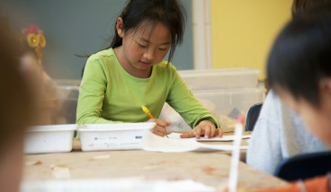A girl writing on paper in a bright room with other children.