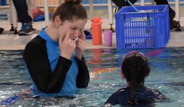 a swimming instructor point to her eyes whilst a child watches, both are in the pool