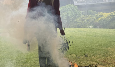 Jillian West conducting Cultural Smoking at an outdoor location. Her gaze is directed towards the fire as she leans forward.